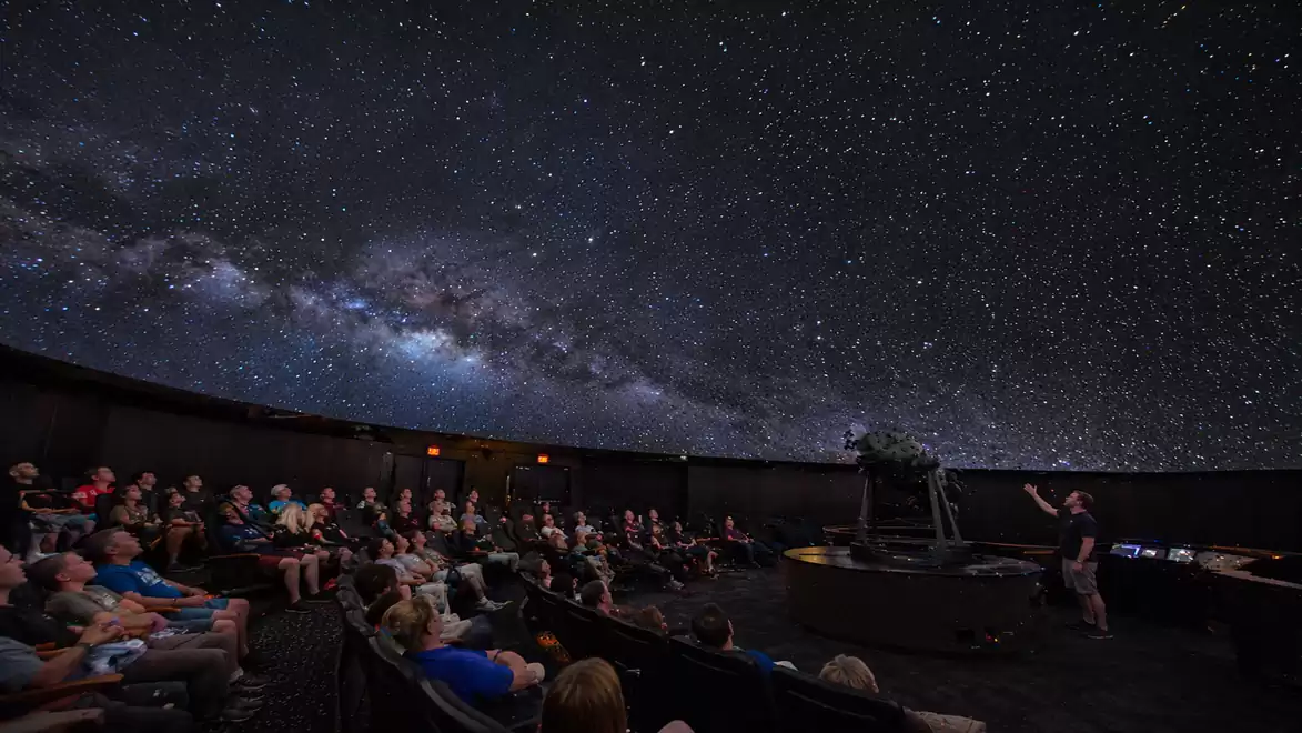 A planetarium with a group of people watching a presentation about the night sky under a projection full of stars and the Milky Way.