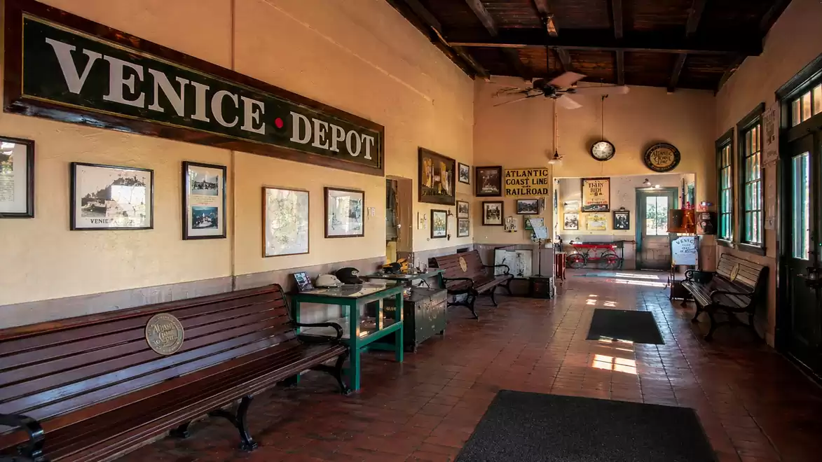 Historical train station waiting room with wooden benches and wall decorations, 'Venice Depot' sign prominently visible.