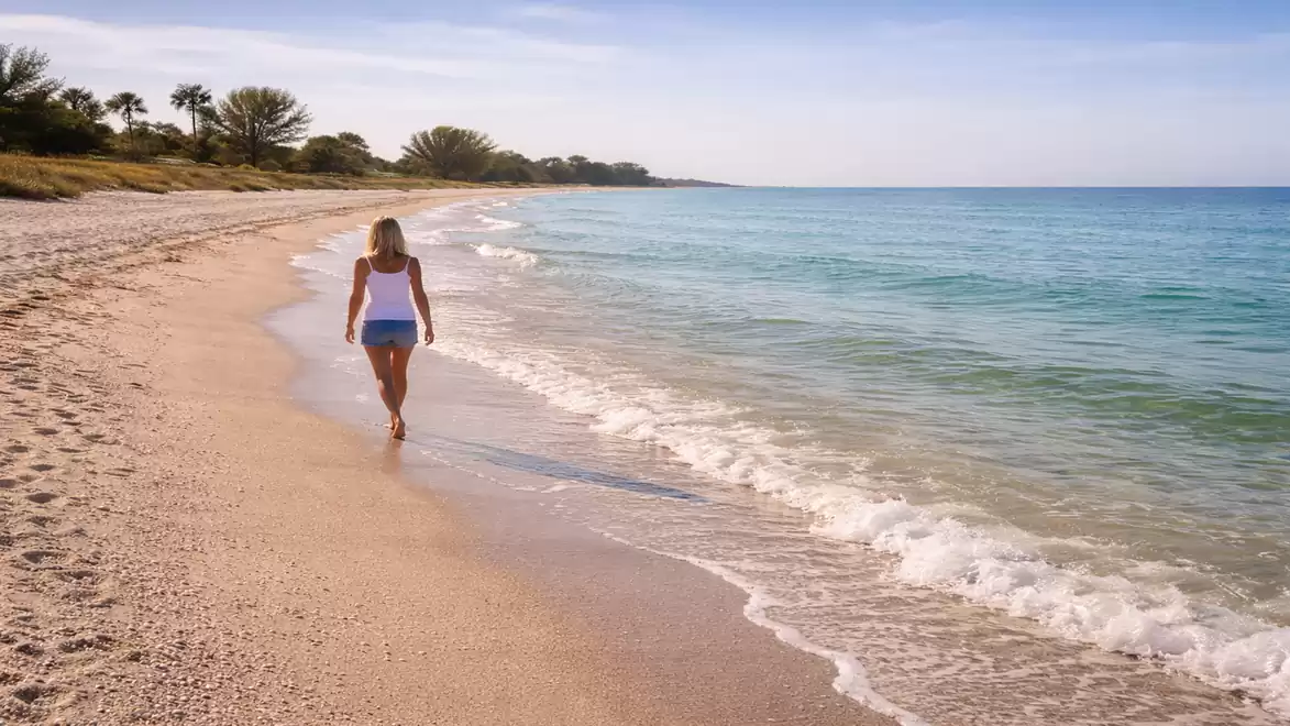 A woman walking along a sandy beach in summer attire, with clear blue water and palm trees in the background.
