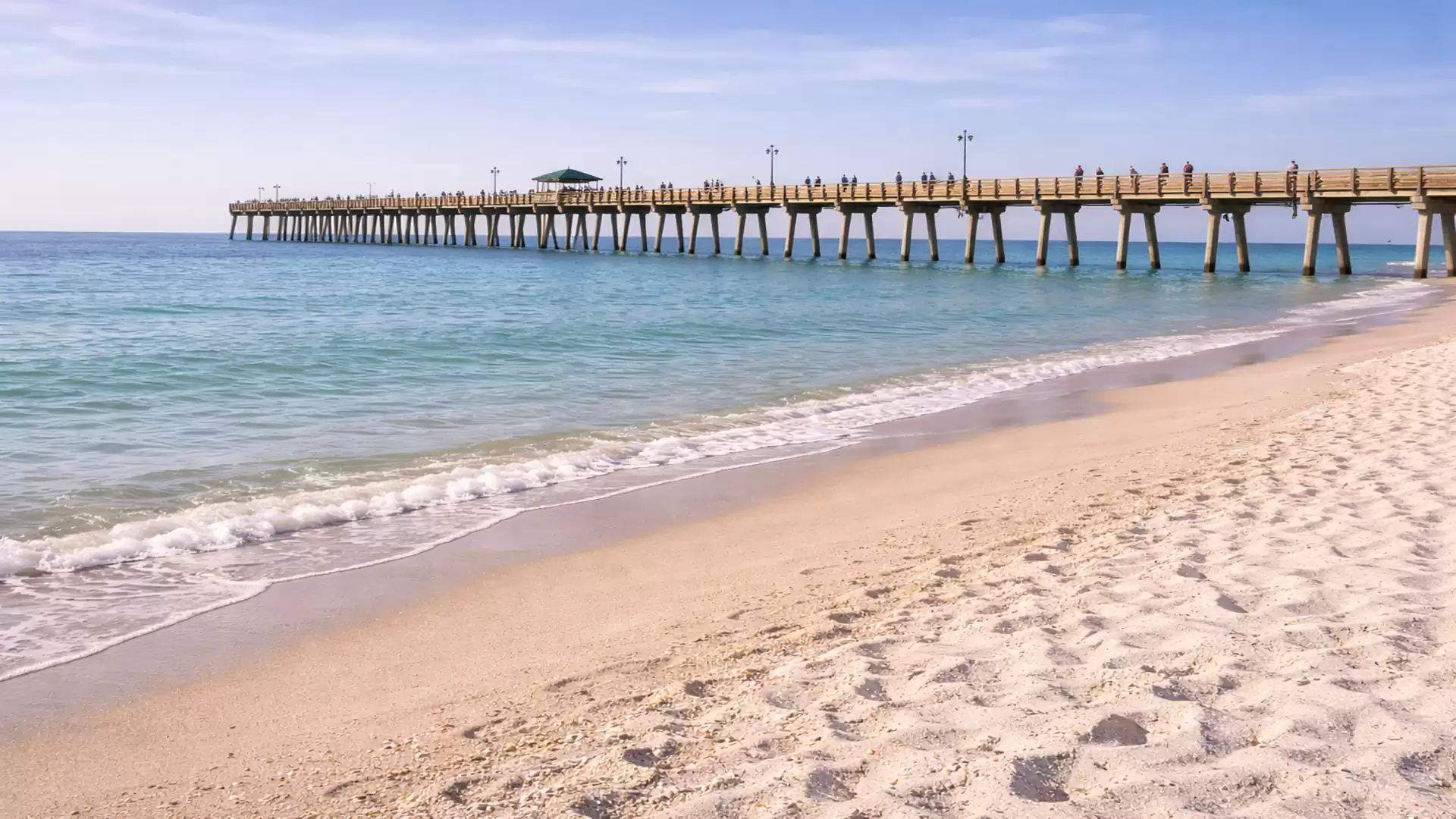 A long wooden pier stretches over the blue sea. The sandy beach in front is empty, the sky clear and sunny.