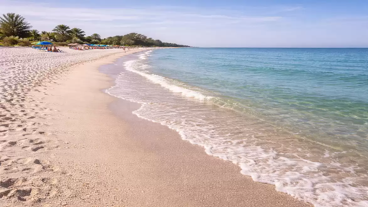 A tranquil sandy beach with clear turquoise water and a few vacationers under parasols in the distance.