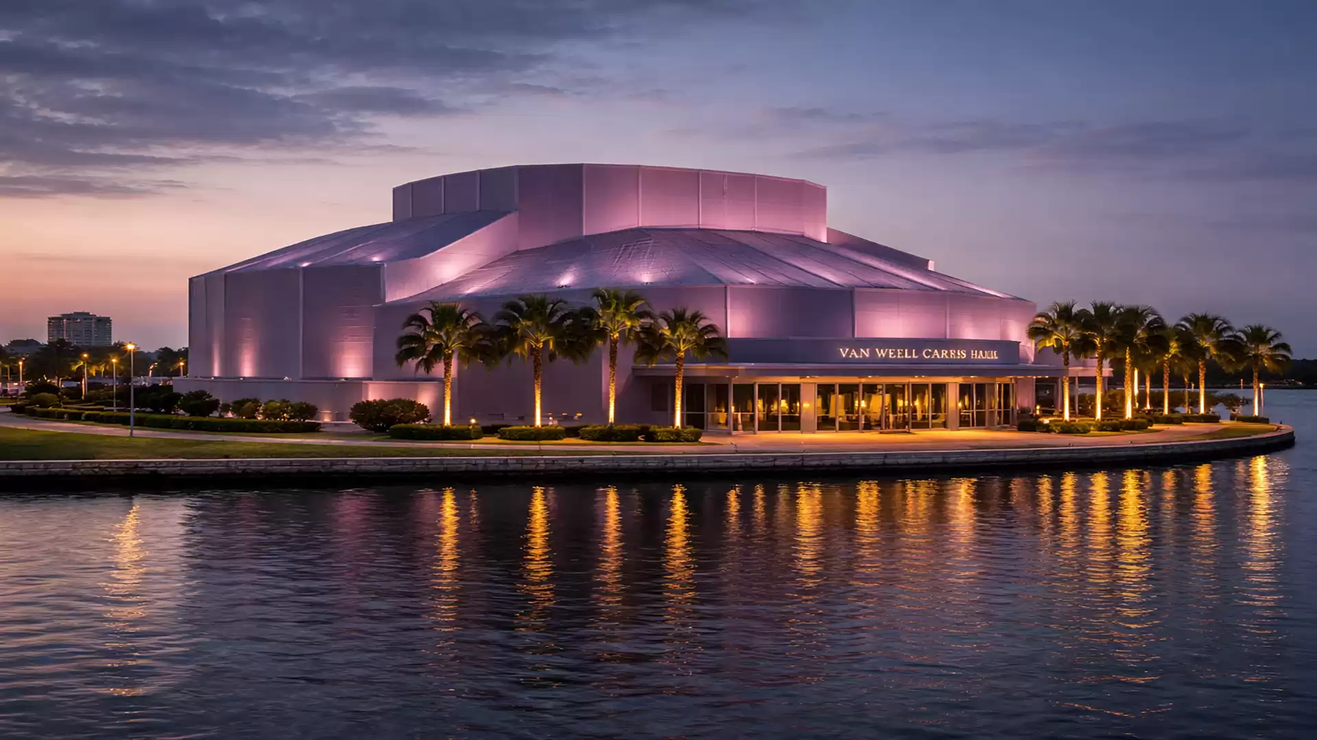 A modern building on the waterfront, illuminated in purple colors, with palm trees in front, surrounded by water at sunset.