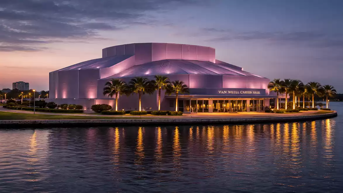 A modern building on the waterfront, illuminated in purple colors, with palm trees in front, surrounded by water at sunset.