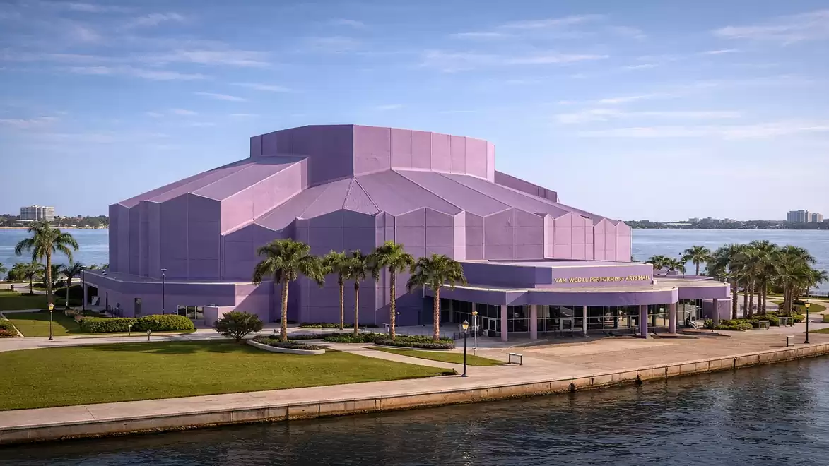 A large, modern building with a purple facade stands by the water, surrounded by palm trees and a blue sky.
