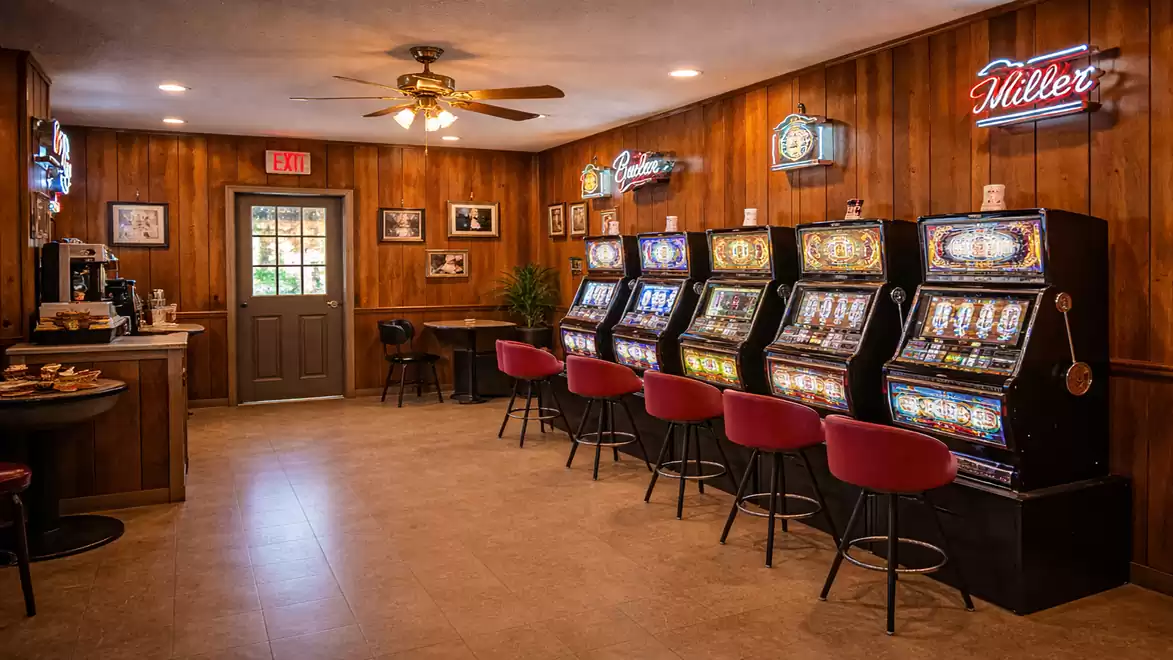 Interior of a gaming room with five slot machines and red stools, wooden walls and a ceiling fan.