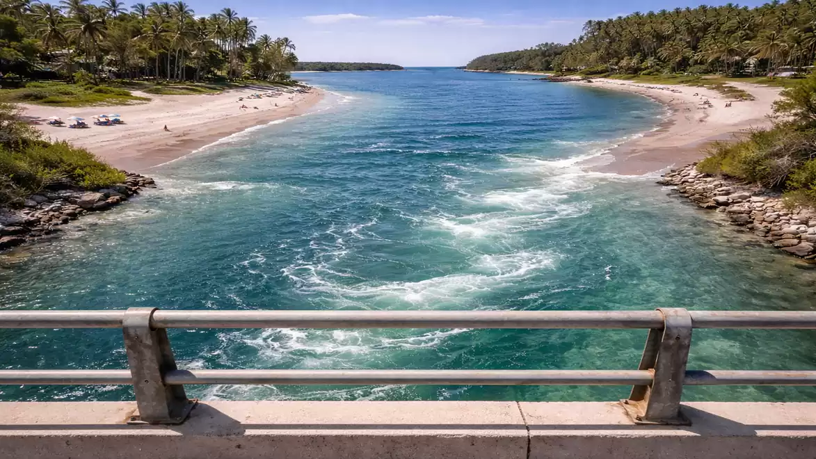 A picturesque view from a bridge overlooking turquoise sea waters flanked by two beaches with palm trees in the distance.