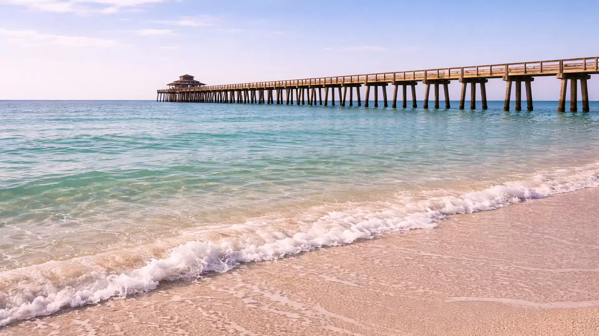 A long wooden pier extends over the blue sea as gentle waves reach the bright sandy beach. The sky is clear and sunny.