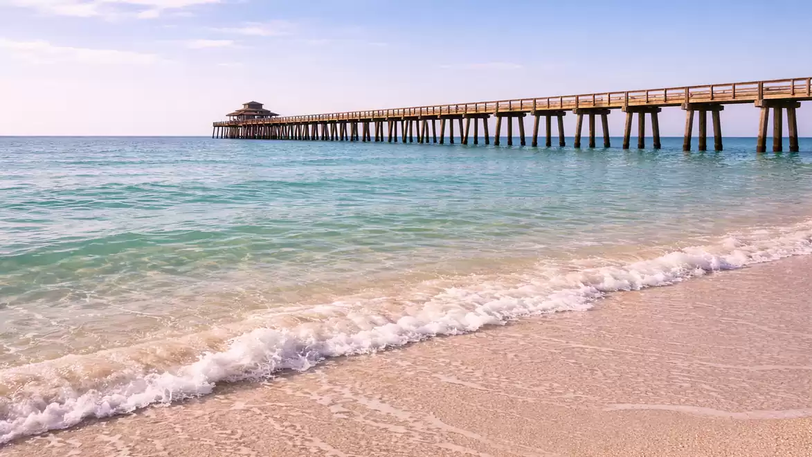 A long wooden pier extends over the blue sea as gentle waves reach the bright sandy beach. The sky is clear and sunny.
