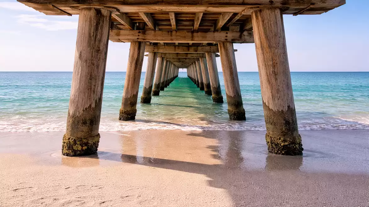 View under a wooden pier towards the turquoise sea, with gentle waves lapping the sandy beach.