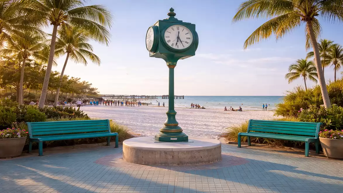 A picturesque beach at sunset featuring a decorative clock in the center, surrounded by green park benches and palm trees.