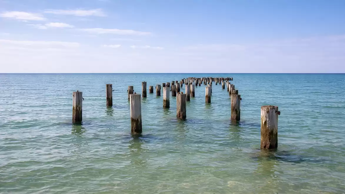 Wooden posts protrude from clear, blue water extending to the horizon, with a clear sky in the background.