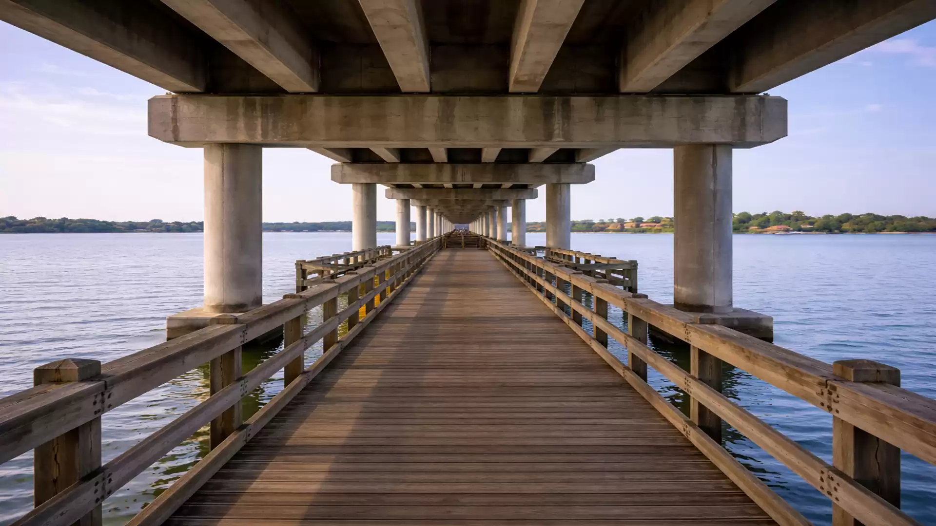 Wooden walkway under a large bridge over a calm body of water with a clear view into the distance.
