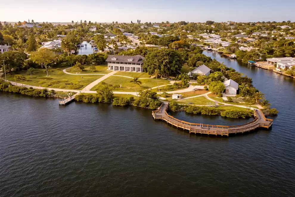 Aerial view of a picturesque house with a large garden by the water, surrounded by lush vegetation and neighboring residential homes in a quiet suburban area.