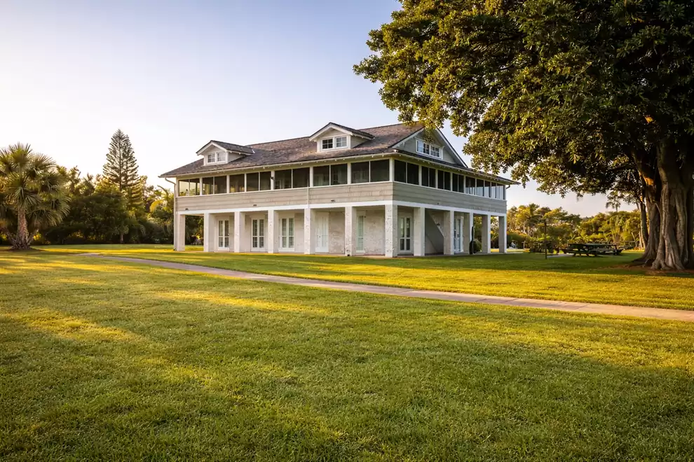 White two-story manor house with a veranda, surrounded by green lawn and large trees at sunset.