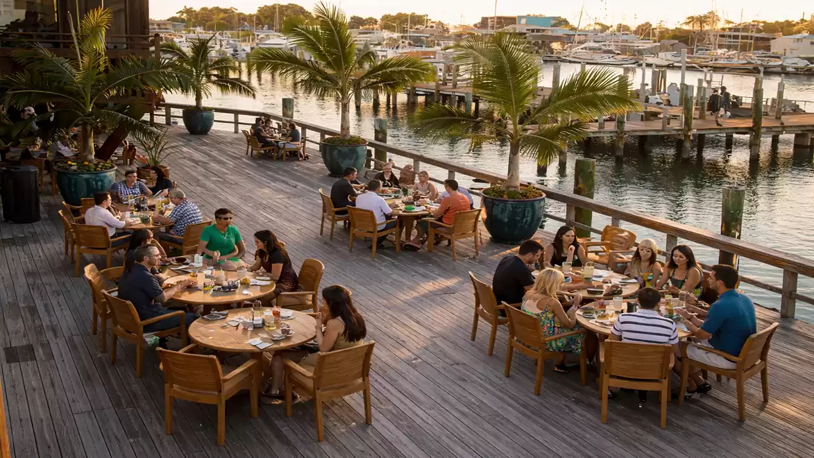 A waterfront restaurant with people dining at wooden tables, surrounded by palm trees and overlooking yachts in the harbor.
