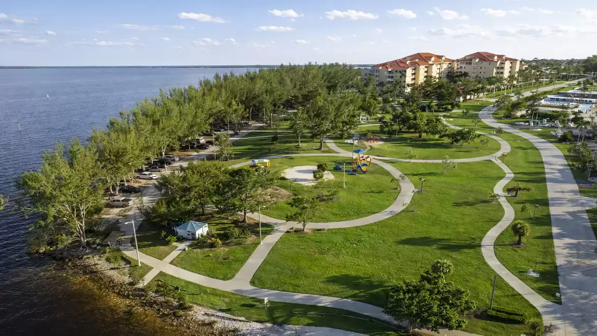 Aerial view of a park along a coastline with green lawns, paths, and a children's playground, surrounded by trees and buildings in the background.