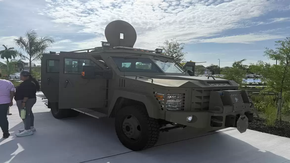 Armored vehicle parked on a sunny day surrounded by a few trees and people standing nearby.