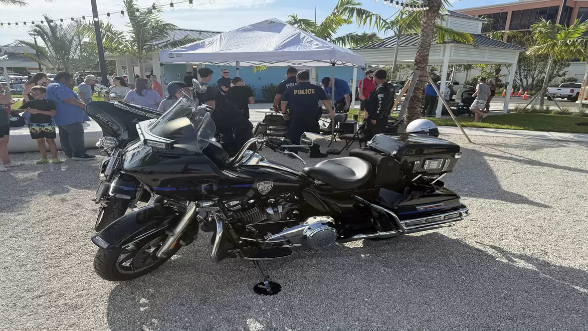 A black police motorcycle is in the foreground, with people under a white tent visible in the background. The scene takes place on a sunny day with palm trees in the background.