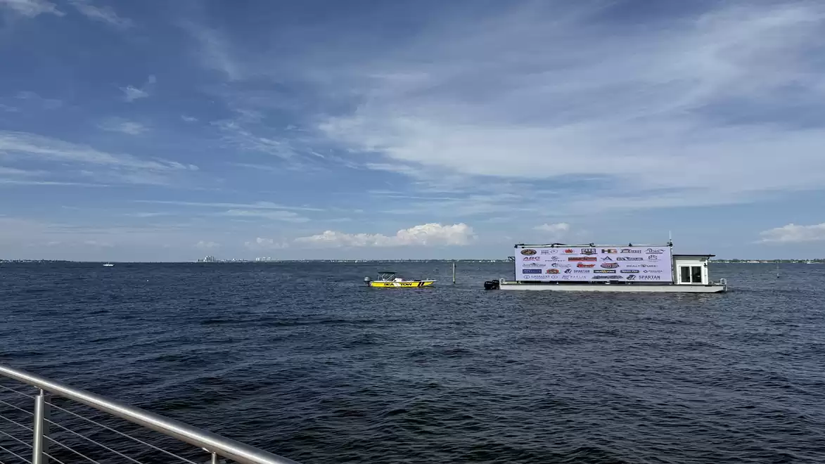 A boat floats on a calm body of water under a clear sky, next to a platform with billboards.