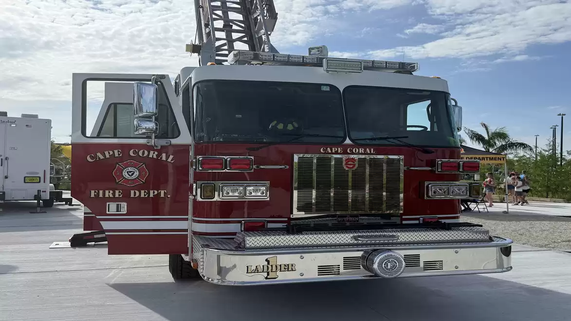 A red fire truck from the Cape Coral Fire Department with an extended ladder, parked on a paved area under a blue cloudy sky.