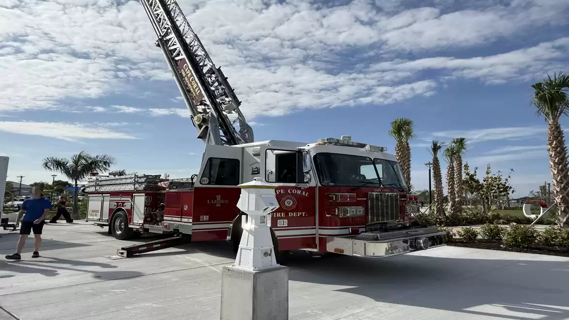 A red fire ladder on a modern fire truck parked on a concrete area with palm trees, under a blue, slightly cloudy sky.