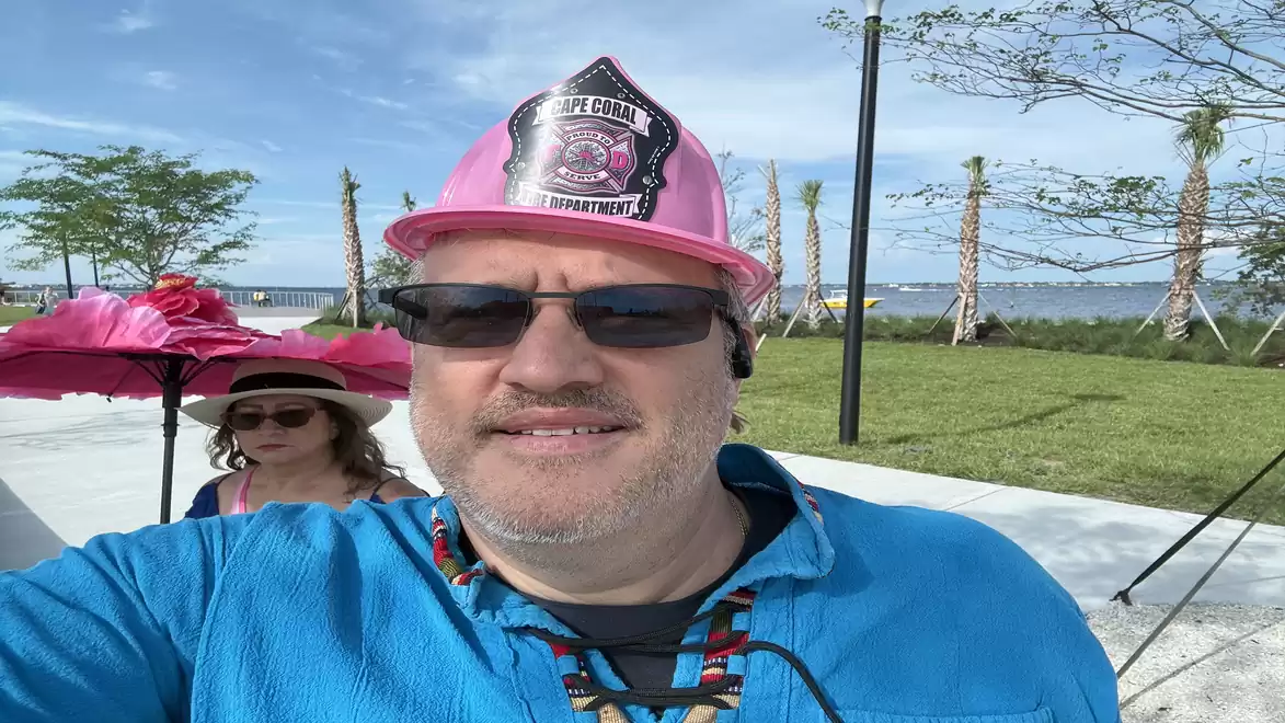 A man wearing sunglasses and a pink helmet stands outdoors on a sidewalk, with a woman in a pink hat and green lawn area in the background.