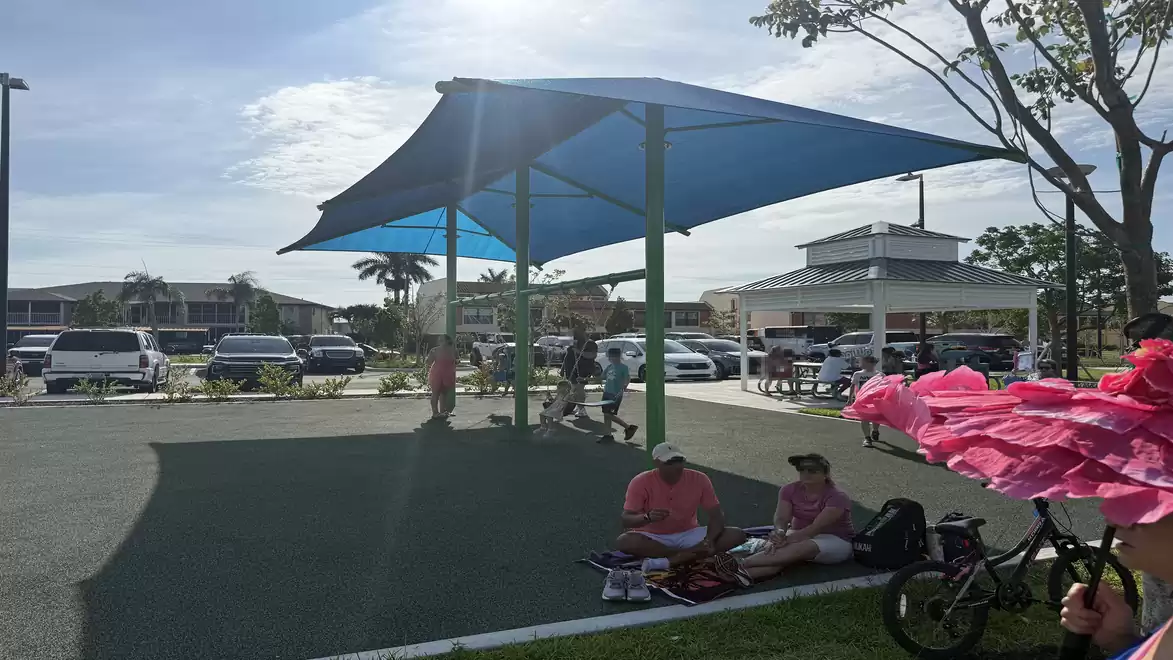 People relaxing in the shade of a large blue canopy on a sunny parking lot. Cars and a white structure are visible in the background.