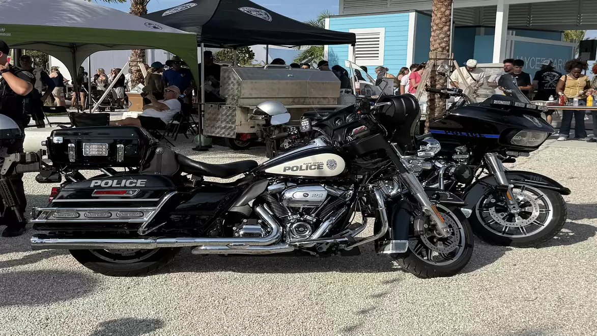 Two police motorcycles are parked in a lot during a public event. Tents and people gathering are visible in the background.