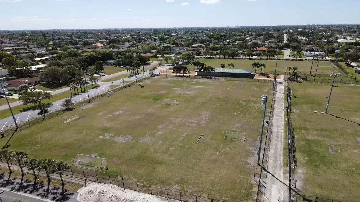 Aerial view of a large empty sports field in an urban setting with surrounding trees and buildings, and a long road on the side.
