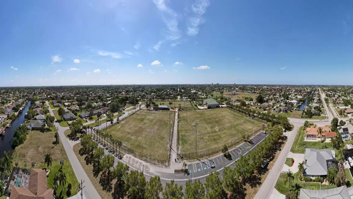 A wide aerial view of a residential area with a large grassy field in the center, surrounded by streets and houses under a clear blue sky.