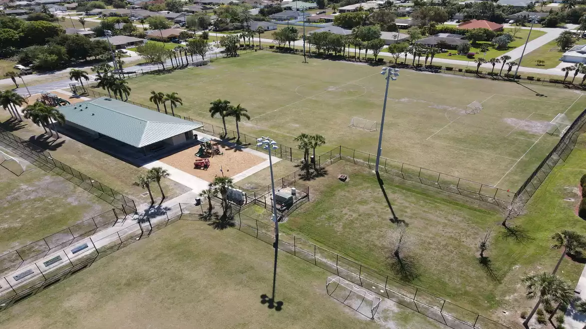 Aerial view of a large green sports field with a central building, palm trees, and surrounding pathways in a residential area.
