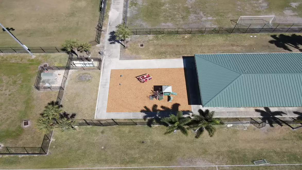 Drone view of a playground with climbing equipment and swings, surrounded by a large park lined with palm trees.