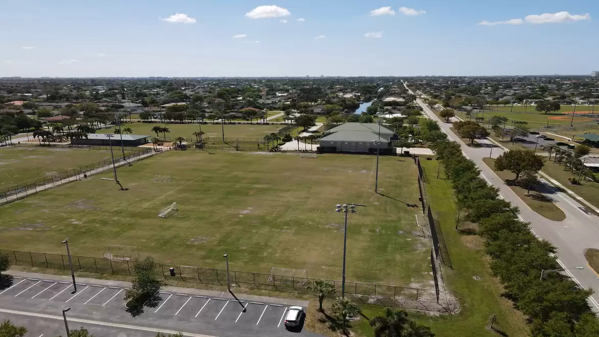 Drone view of a large sports field surrounded by roads and buildings under a blue sky.