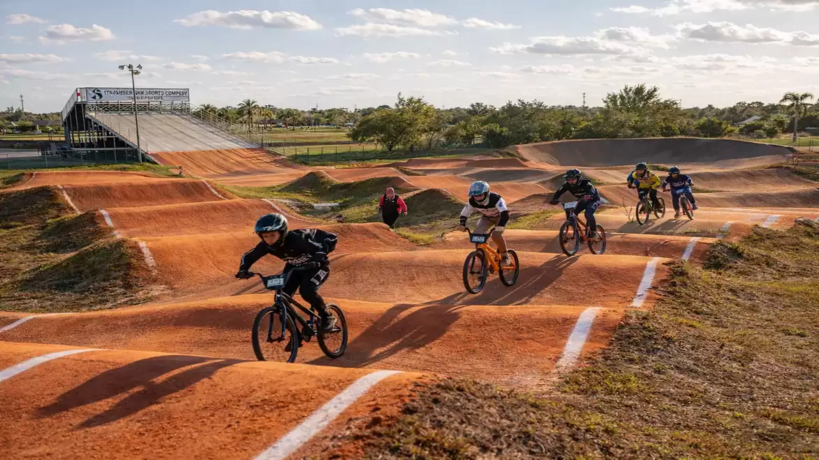 Five BMX riders in full protective gear are racing on a winding dirt track on a sunny day.