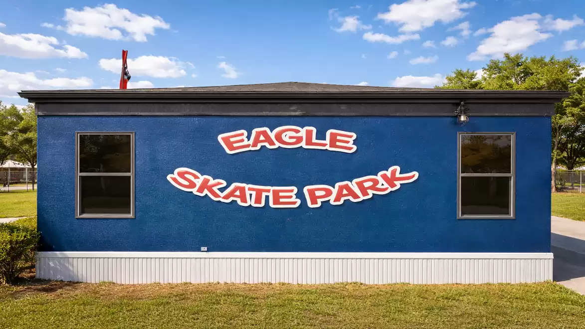 Blue building with the sign 'Eagle Skate Park' in large red letters, surrounded by grass and trees under a blue sky.