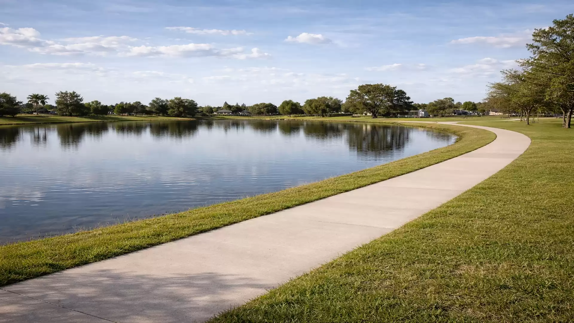 A peaceful lake under a clear sky with a curved walkway along the shore, surrounded by green landscape.