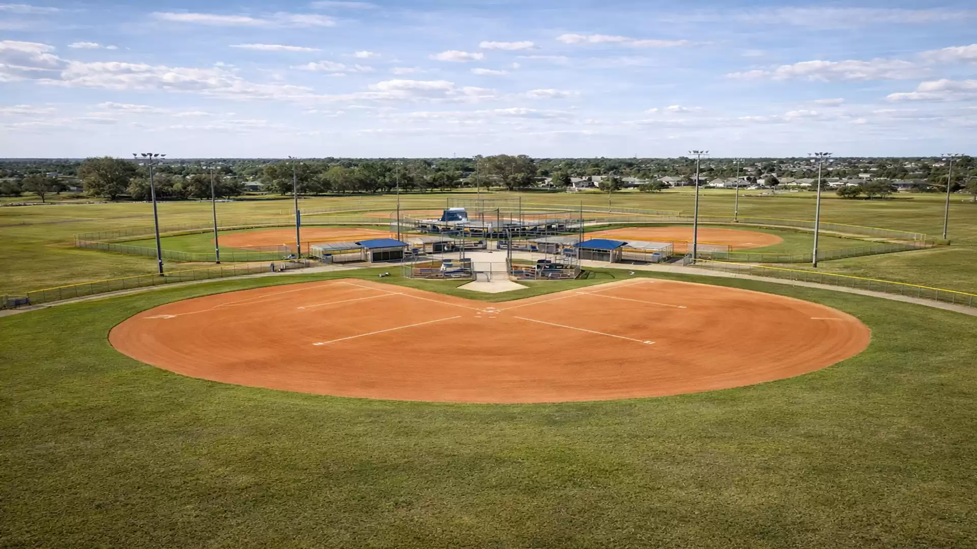 Aerial view of an empty baseball field in a vast green area under a blue sky with a few clouds.