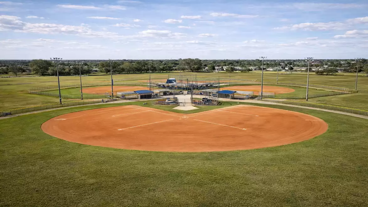 Aerial view of an empty baseball field in a vast green area under a blue sky with a few clouds.