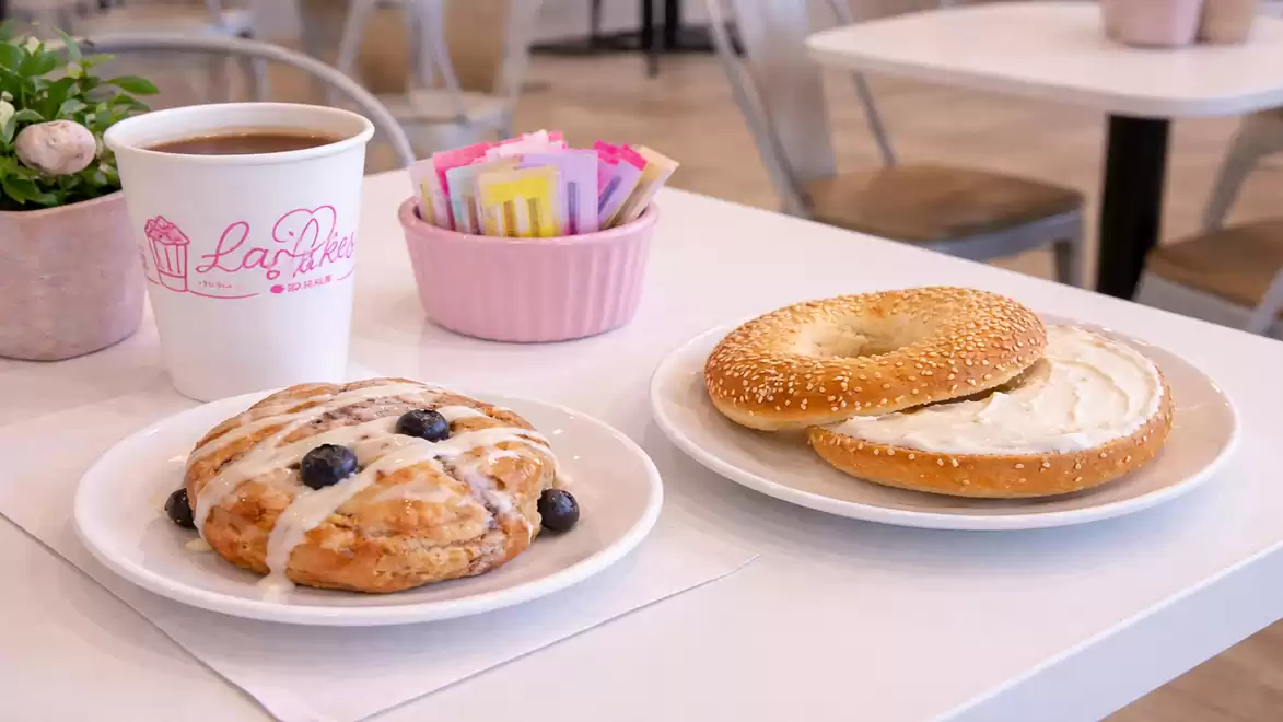 A table in a café with a hot beverage in a white cup, a blueberry muffin on a plate, and a bagel with spread.