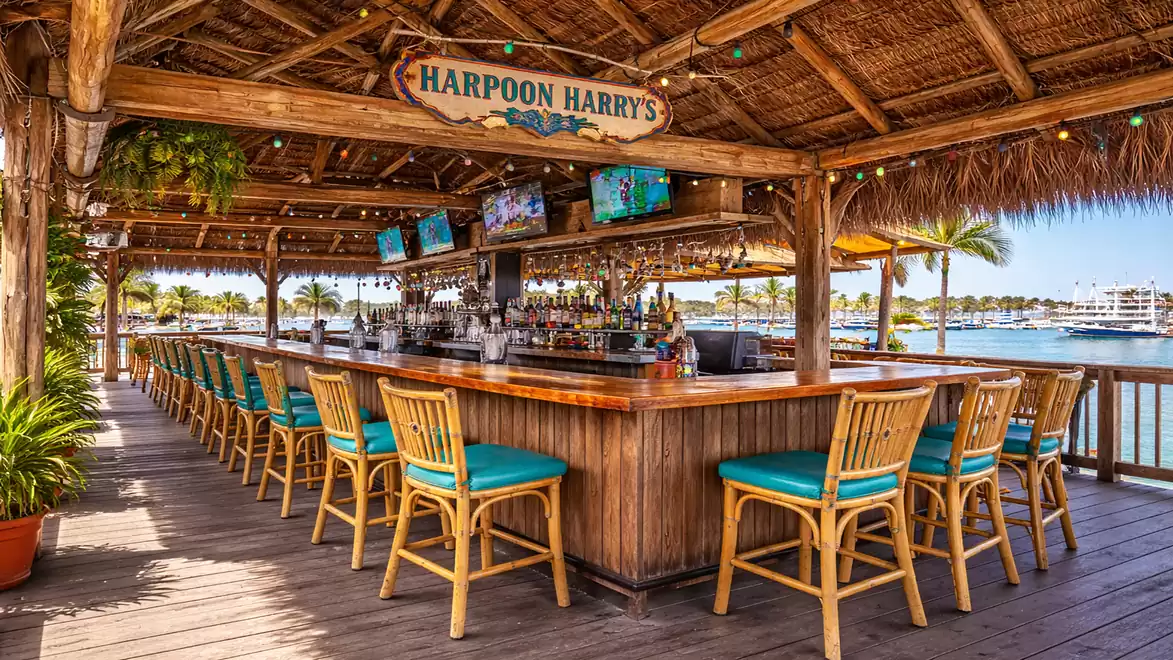 A beach bar with a wooden structure and thatched roof, surrounded by wooden bar stools with turquoise cushions. In the background, palm trees and the sea are visible.