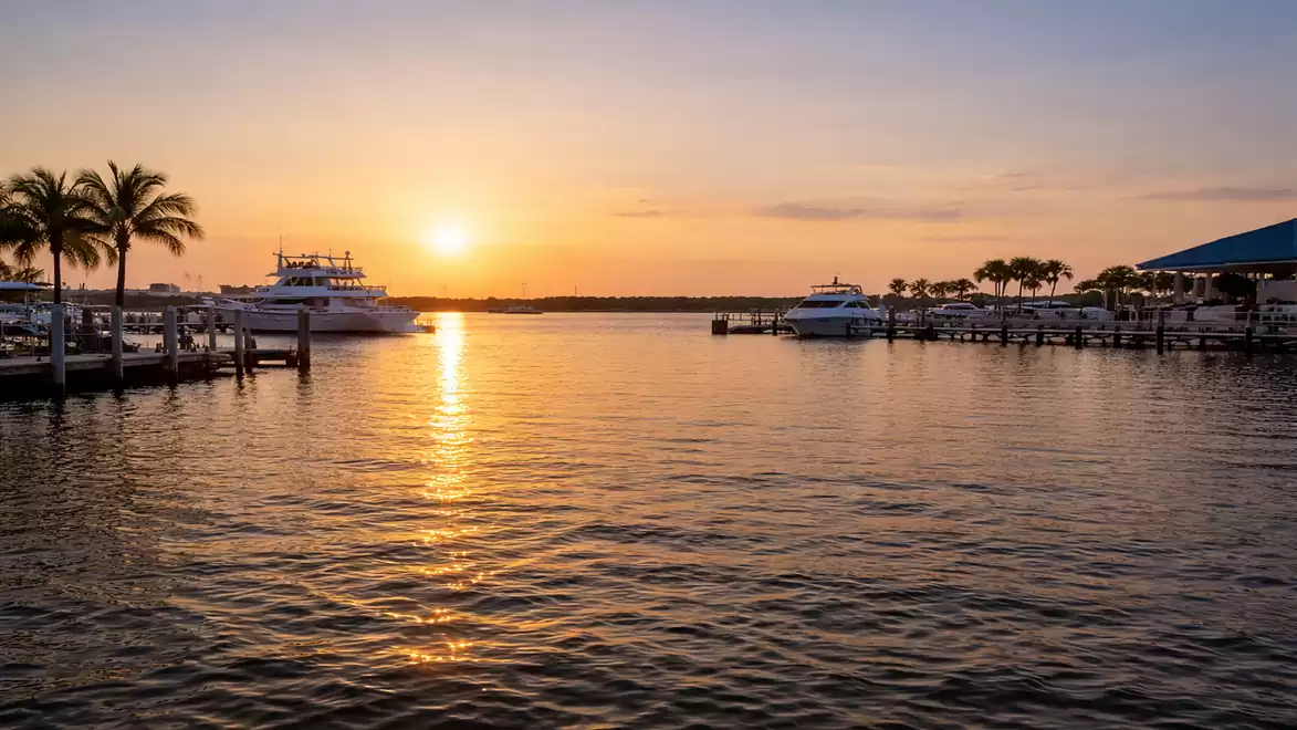 A picturesque sunset over a tranquil harbor, with palm trees, boats, and a vividly colorful sky.