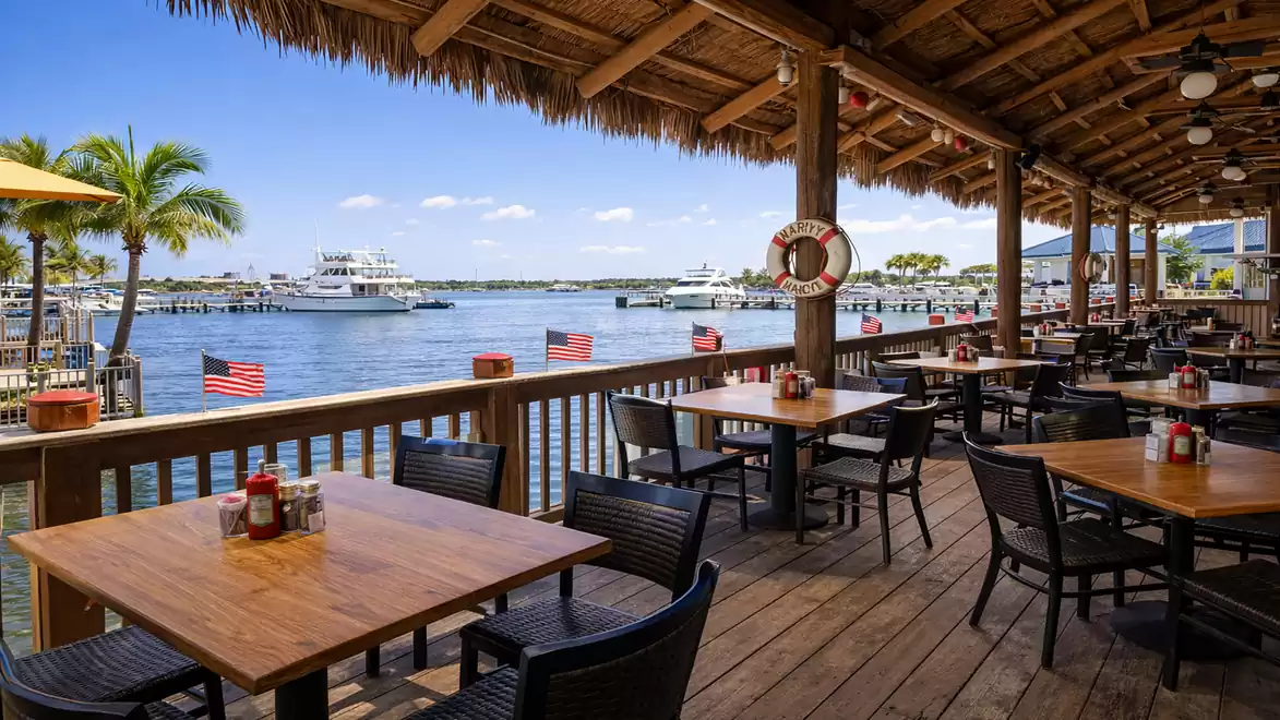 A restaurant with a wooden deck overlooking a harbor. Several yachts and boats are visible in the background. The tables and chairs are empty, and American flags are placed along the railing.