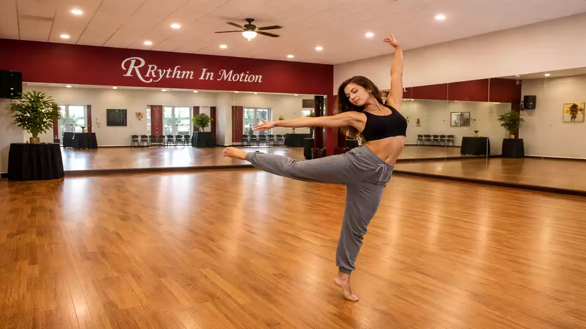 A woman dances in a spacious, bright dance studio in front of a mirror. She is wearing a black top and gray sweatpants.