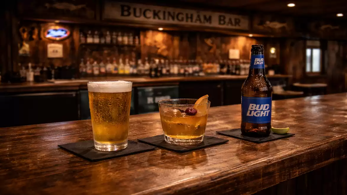 Three drinks on a rustic bar: a glass of beer with foam, a cocktail glass with an orange slice and cherry, and a Bud Light bottle with a lime slice.