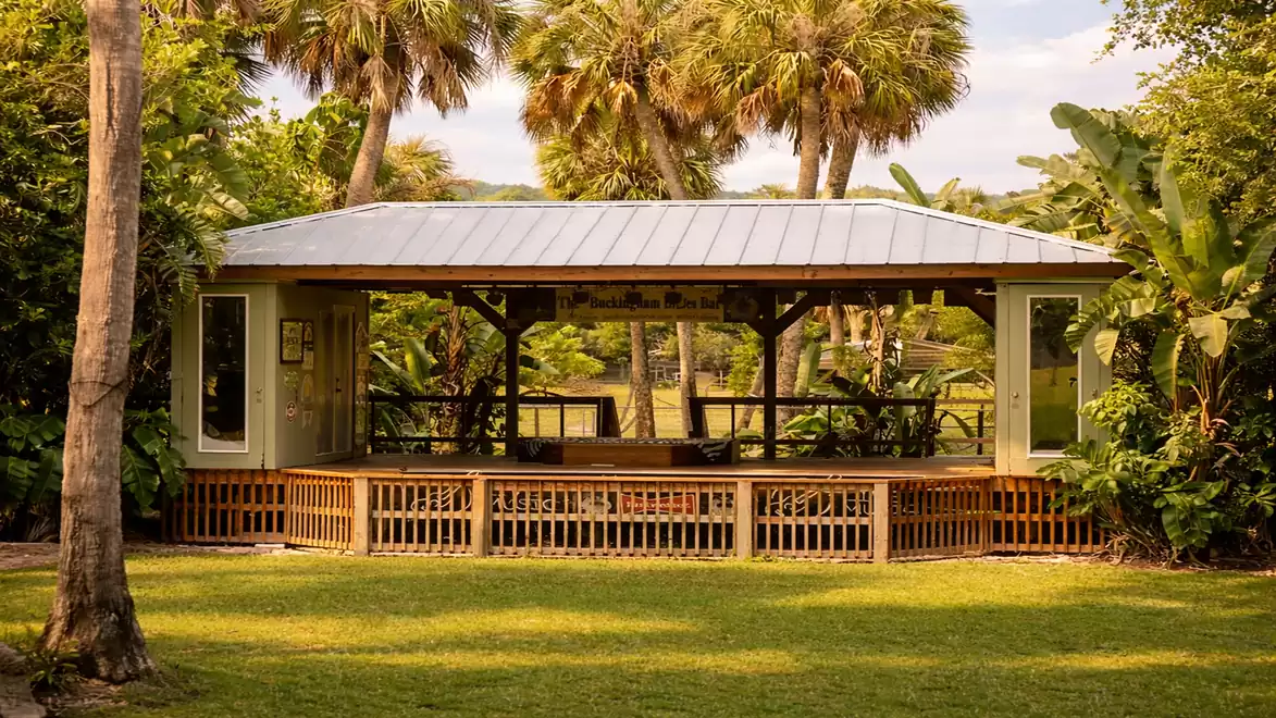 A spacious pavilion in a green garden with palm trees in the background. The pavilion has a white roof and is surrounded by lush vegetation.