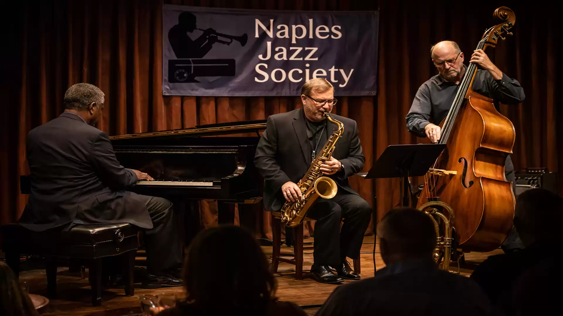 A jazz band performs on stage. A pianist, saxophonist, and double bassist are visible. A banner reading 'Naples Jazz Society' hangs in the background.