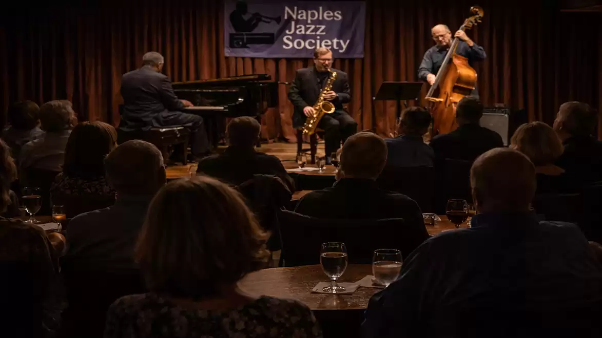 A jazz band performs in front of an audience in a club. A pianist, a saxophonist, and a double bassist are playing under a Naples Jazz Society banner.