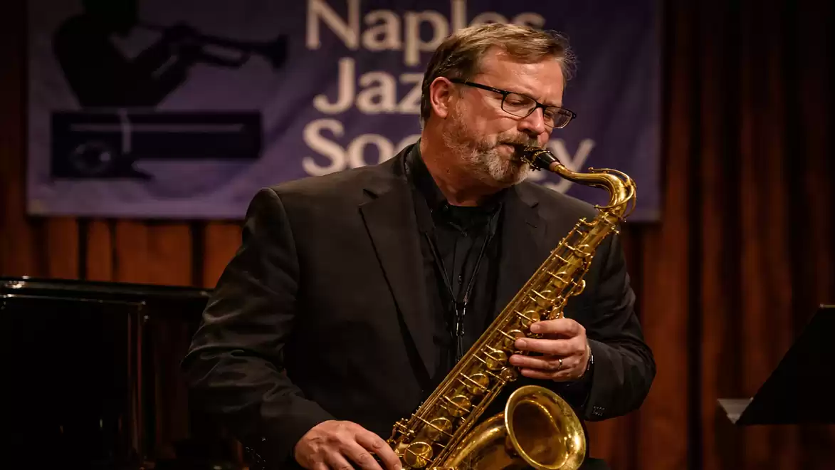 A musician plays saxophone on stage, with a Naples Jazz Society banner visible in the background.