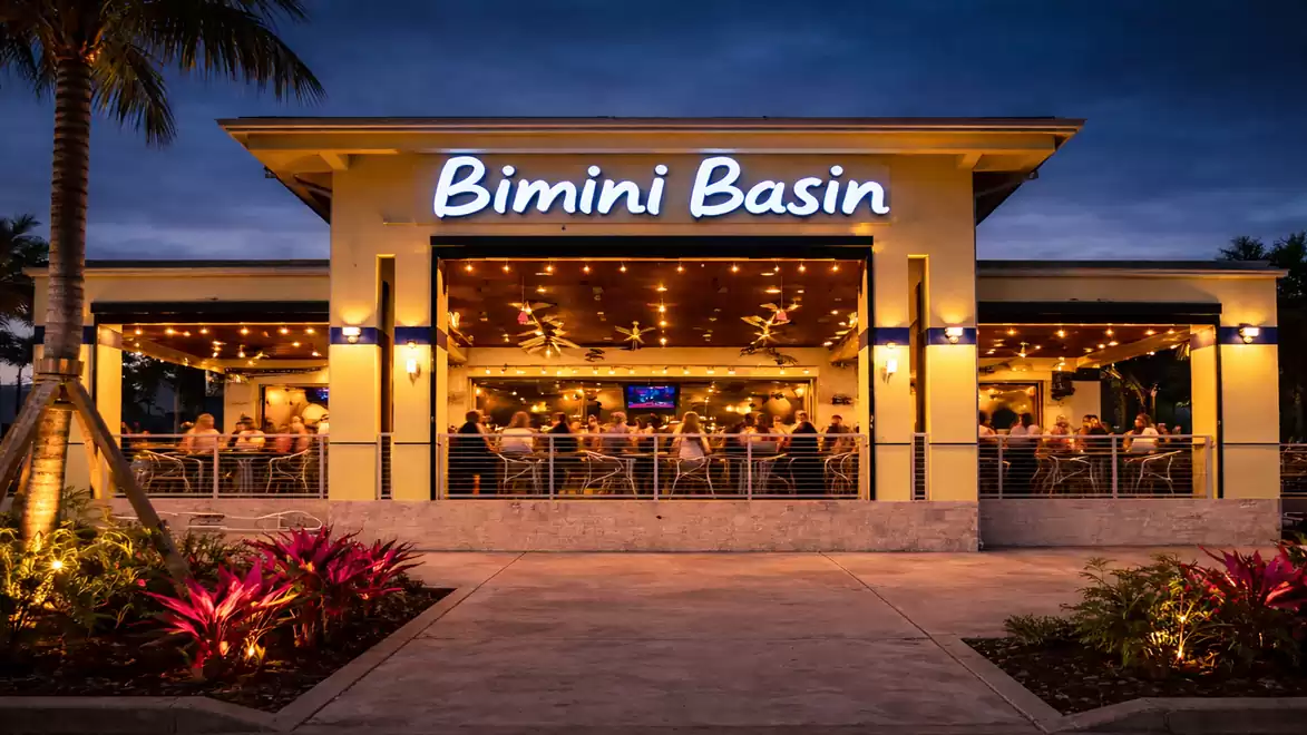 Evening view of a lively restaurant named Bimini Basin with a lit outdoor terrace and palm trees in the background.