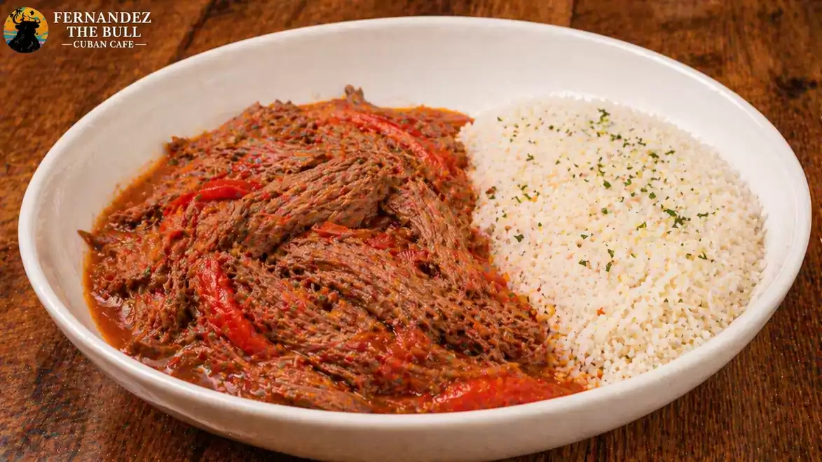 A plate of Ropa Vieja, a traditional Cuban dish consisting of shredded beef in a tomato sauce with red pepper strips, served with a side of rice sprinkled with herbs.